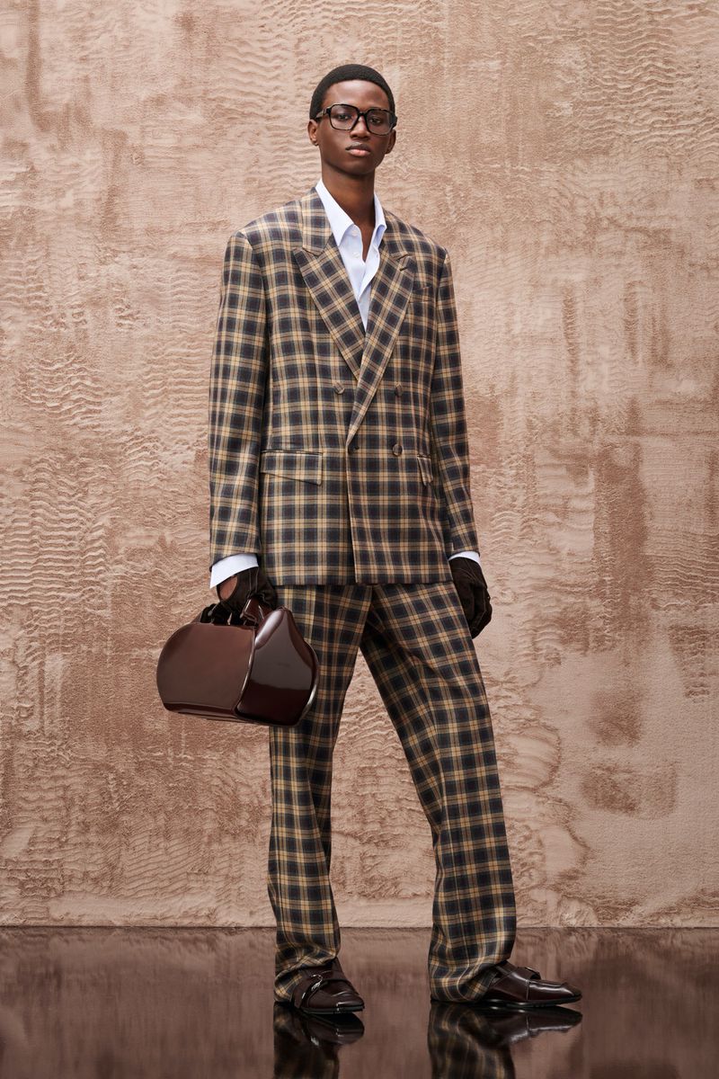 A Black male model in a checkered suit poses in front of a neutral background, carrying a structured handbag and wearing glasses.