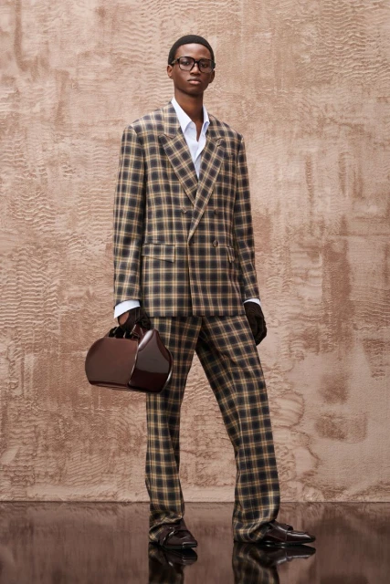 A Black male model in a checkered suit poses in front of a neutral background, carrying a structured handbag and wearing glasses.
