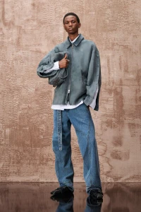 Model standing against a textured brown wall wearing a blue jacket, white shirt, and wide blue jeans while holding a matching bag