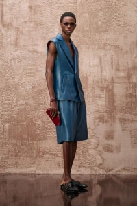 Model wearing a sleeveless blue leather jacket and matching shorts holding a red clutch against a textured wall.
