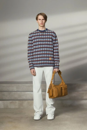 Male model in a patterned knit sweater and white trousers holding a tan leather shoulder bag against a set-dressed studio background