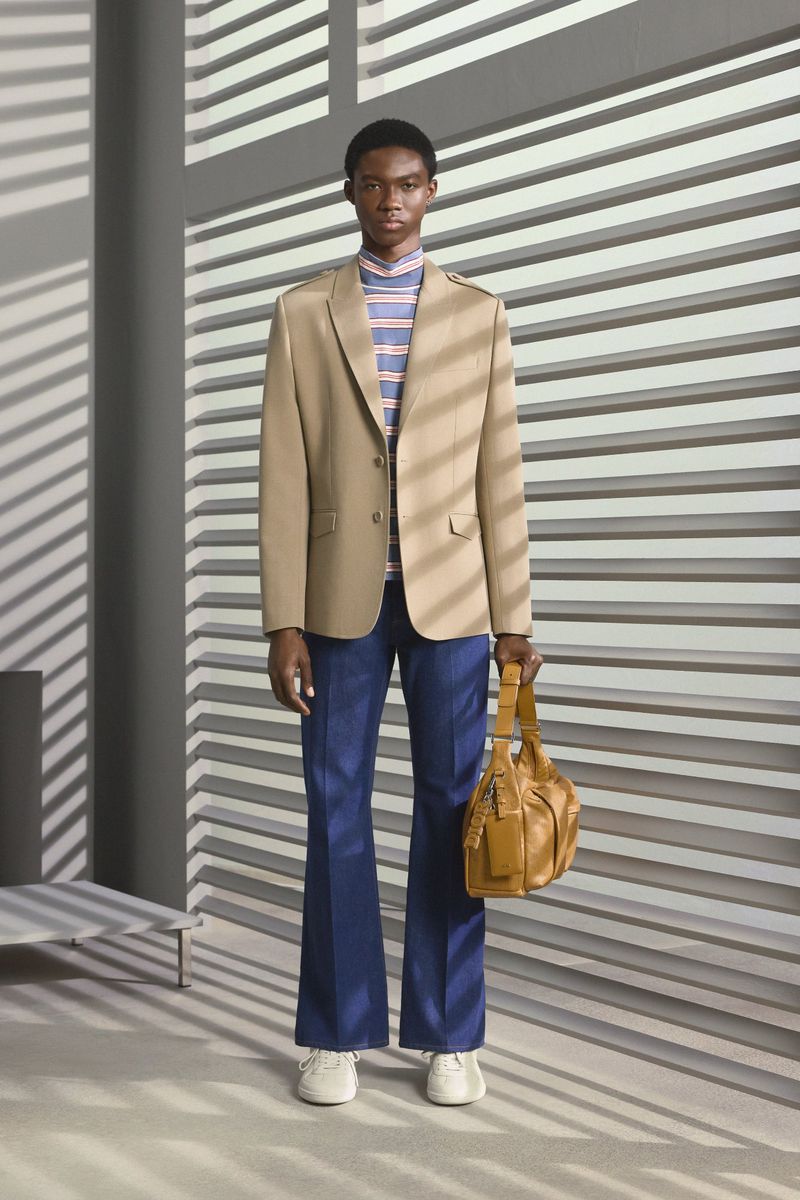 A male fashion model poses indoors wearing a blazer, striped turtleneck, jeans, and carrying a leather bag in a full shot.