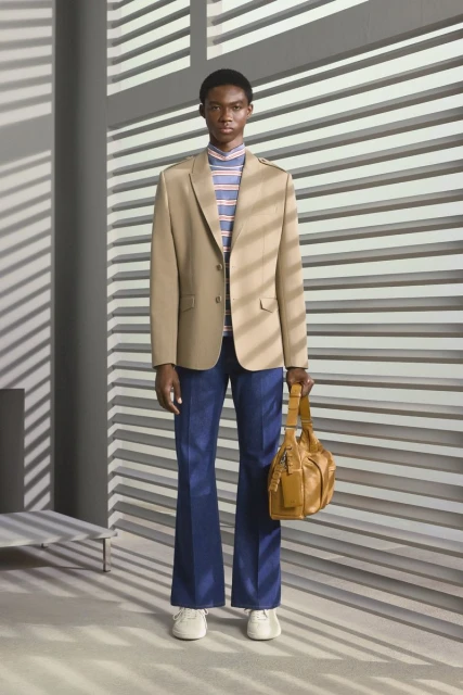 A male fashion model poses indoors wearing a blazer, striped turtleneck, jeans, and carrying a leather bag in a full shot.