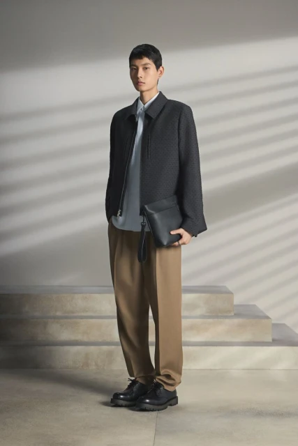 Portrait of a young man in a dark jacket and brown pants, holding a black clutch, standing against a neutral background with stairs.