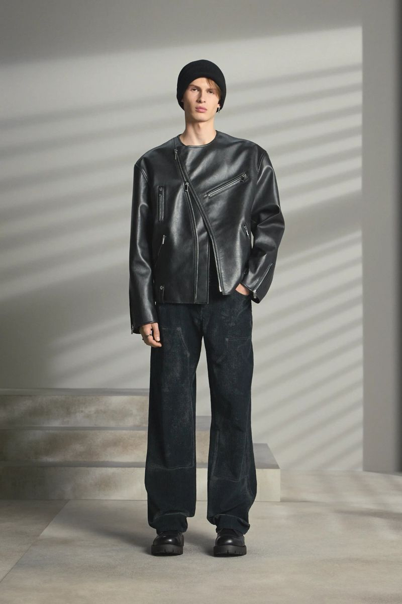 Man stands in a photo studio wearing a black leather jacket, black beanie, and dark trousers, illuminated by soft, diffused lighting.