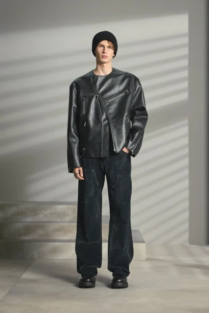 Man stands in a photo studio wearing a black leather jacket, black beanie, and dark trousers, illuminated by soft, diffused lighting.