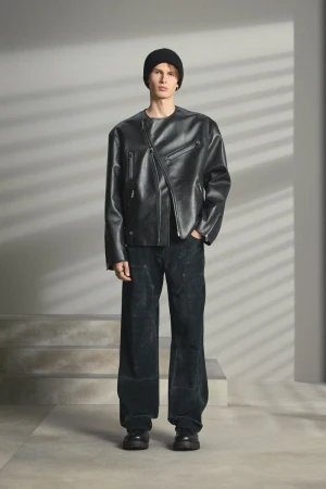 Male model in a black leather jacket and beanie standing against a gray wall with horizontal light stripes.