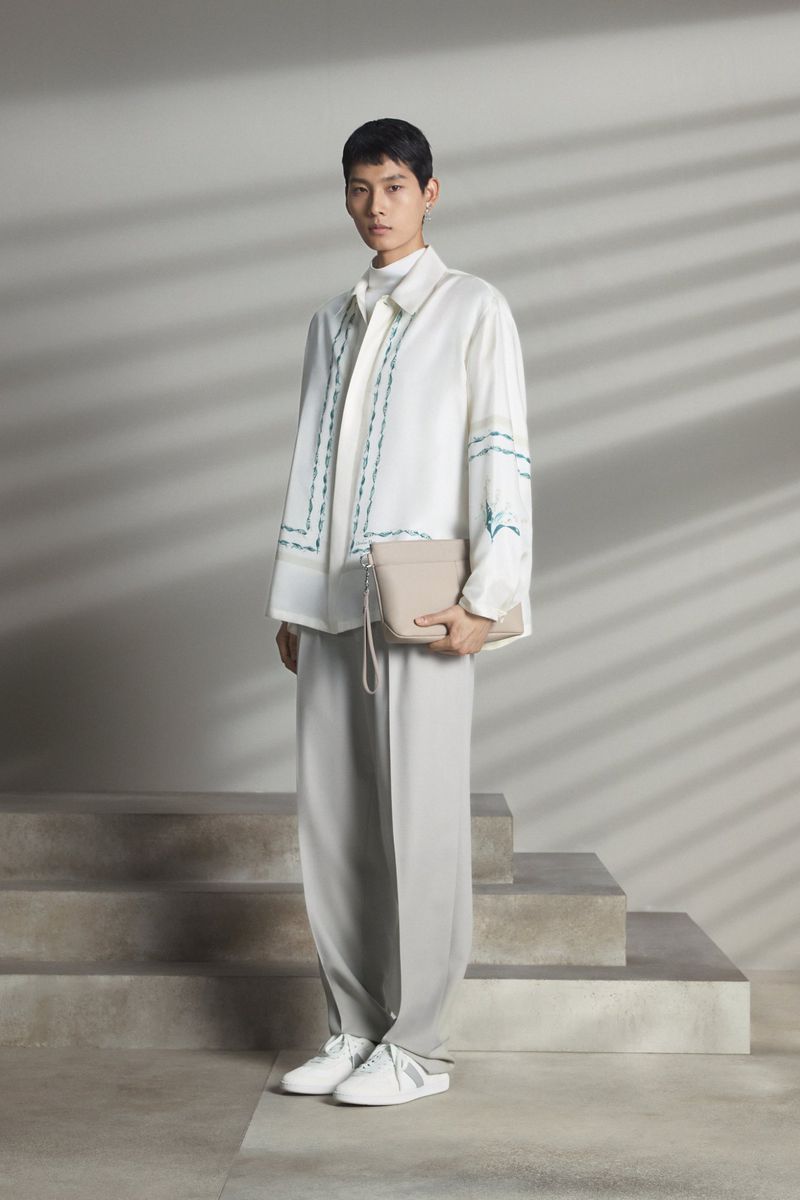 Fashionable adult Asian man in a white shirt and grey pants holding a purse, standing against a grey wall with shadow patterns.