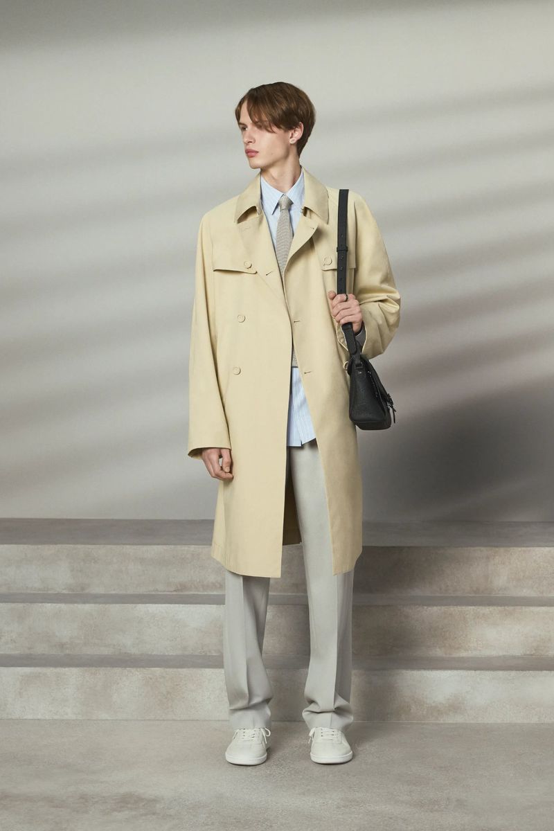 A slender man in a light trench coat and grey pants poses on steps, wearing a blue shirt, tie, white sneakers, and carrying a black bag.