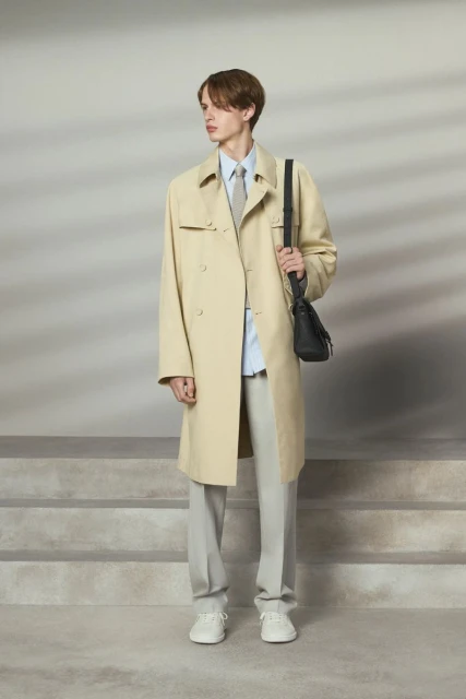 A slender man in a light trench coat and grey pants poses on steps, wearing a blue shirt, tie, white sneakers, and carrying a black bag.