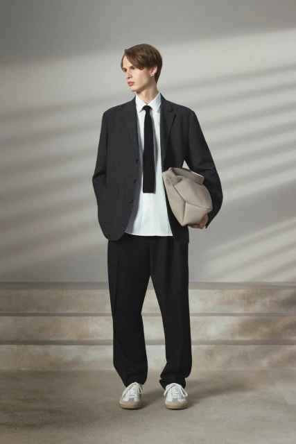 A slender man in a suit stands on stairs, holding a bag, with light creating window shadow patterns on the neutral wall behind.
