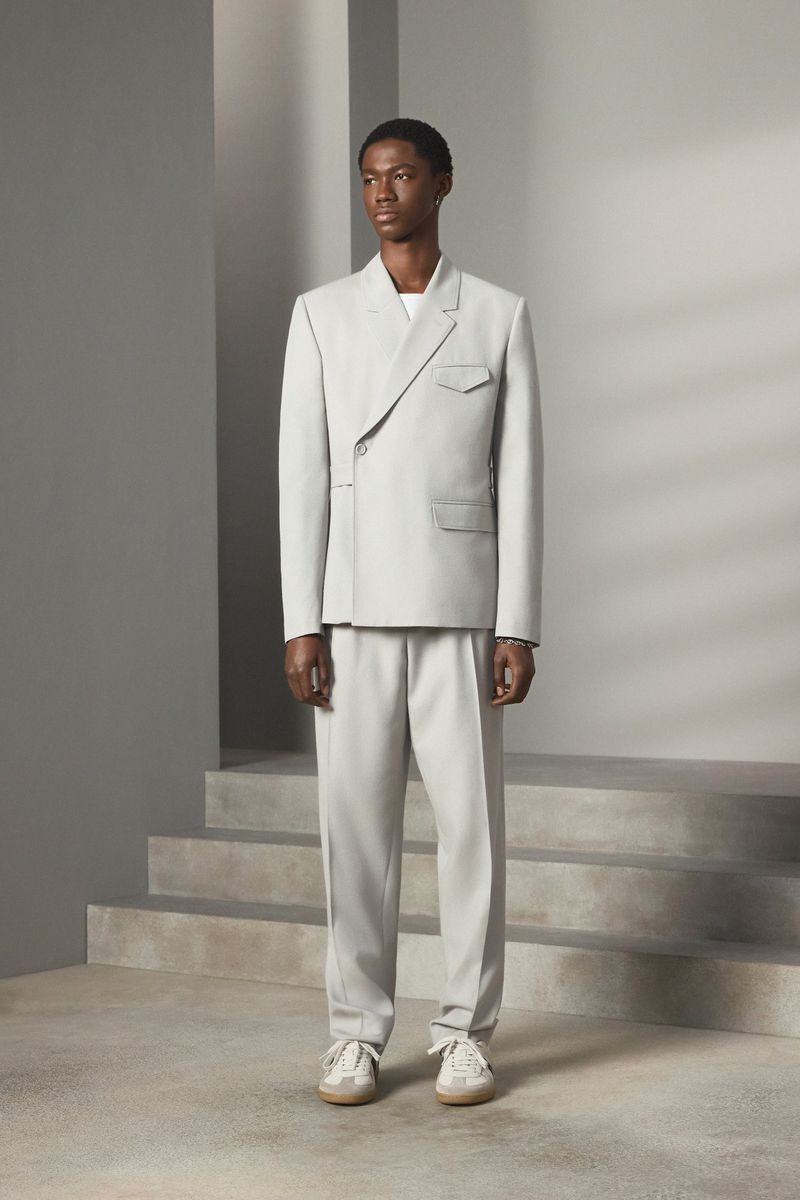 Dark-skinned male fashion model stands in a light grey suit, light trainers, in a studio with stair-like architectural details.