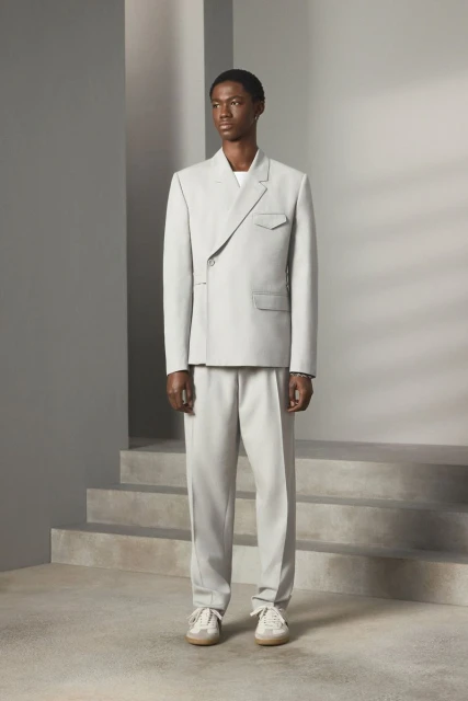 Dark-skinned male fashion model stands in a light grey suit, light trainers, in a studio with stair-like architectural details.