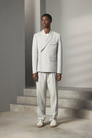 Dark-skinned male fashion model stands in a light grey suit, light trainers, in a studio with stair-like architectural details.