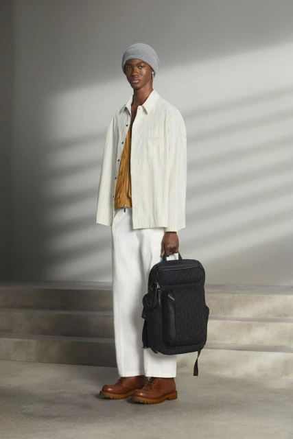 Male fashion model poses in an off-white button-down shirt, white pants, brown shoes, and grey beanie, holding a black backpack.