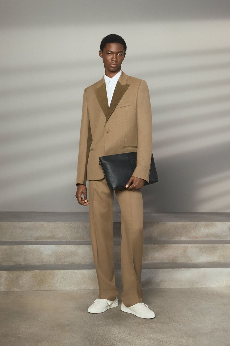 Male fashion model in a tan suit and white sneakers holding a black clutch, posing indoors against a neutral backdrop with soft lighting.