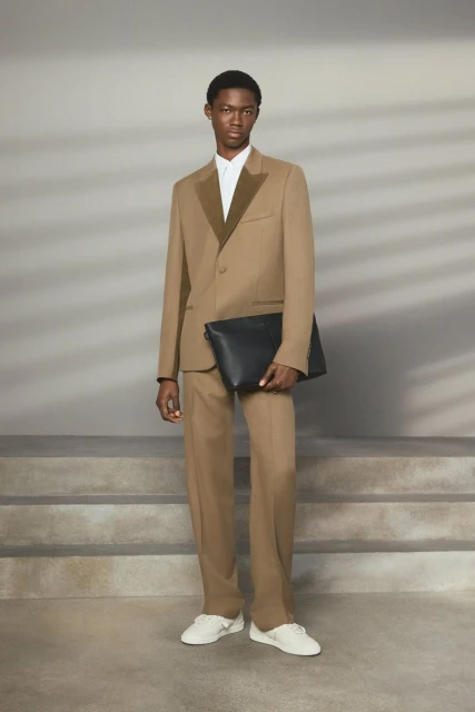 Male fashion model in a tan suit and white sneakers holding a black clutch, posing indoors against a neutral backdrop with soft lighting.