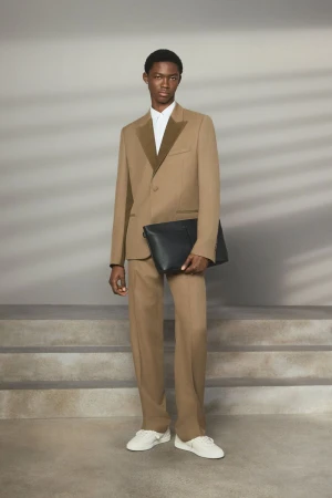 Male fashion model in a tan suit and white sneakers holding a black clutch, posing indoors against a neutral backdrop with soft lighting.