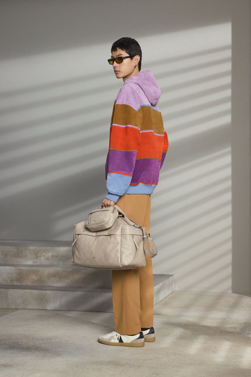 Side view of a person wearing sunglasses, a striped hoodie, and tan pants holding a beige duffel bag with small pouches attached.