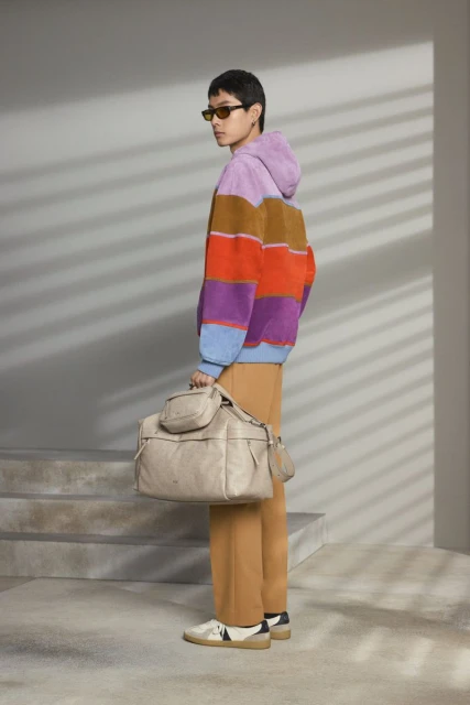 Side view of a person wearing sunglasses, a striped hoodie, and tan pants holding a beige duffel bag with small pouches attached.