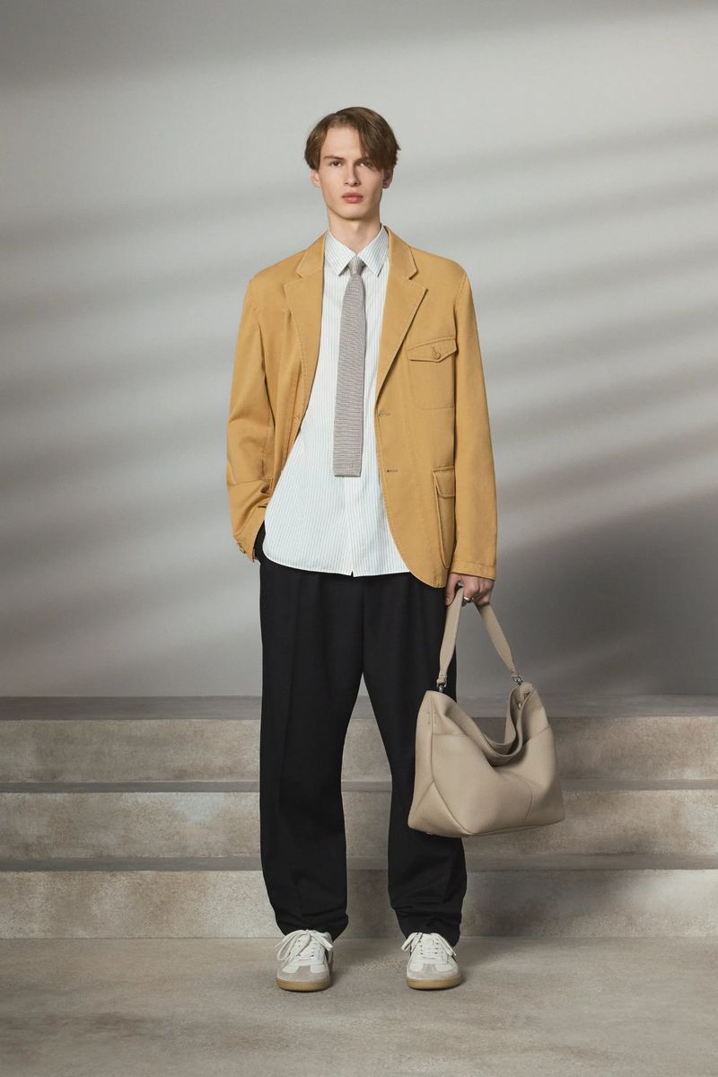 A slender man poses on concrete steps in a relaxed, modern style with a tan jacket, white shirt, black pants, tie, shoulder bag, and sneakers.