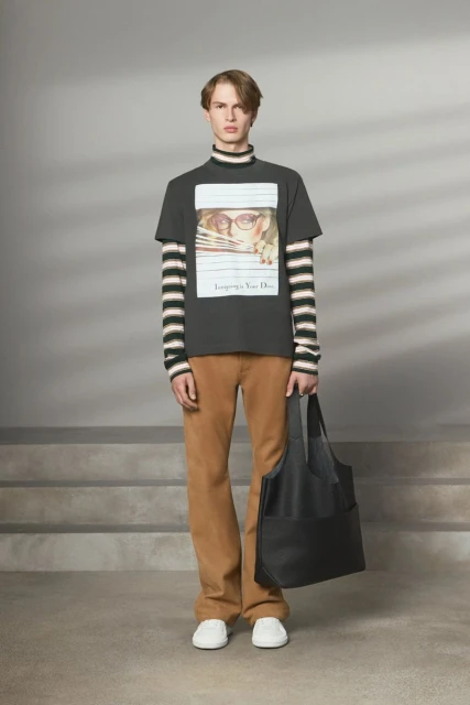 Male fashion model posing indoors wearing a printed t-shirt, brown pants, and a black tote bag in a studio setting.