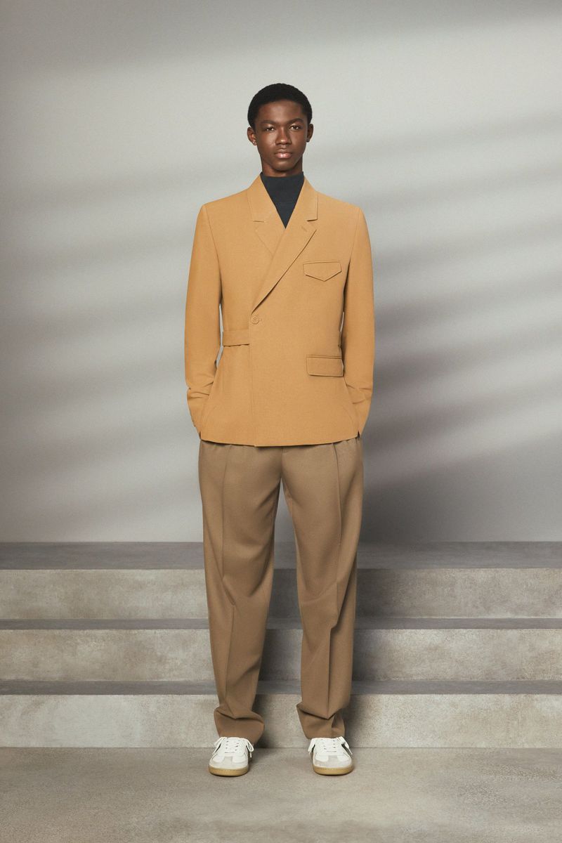 An adult Black male fashion model poses in a caramel suit with a black mock turtleneck and tan pants on a gray background.