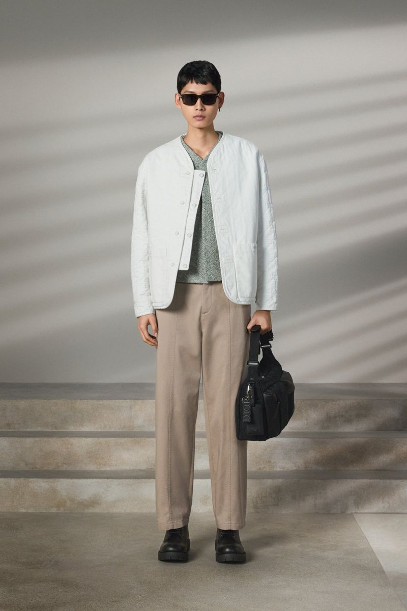 Model wearing a light grey quilted jacket, brown trousers, and sunglasses while holding a black shoulder bag.