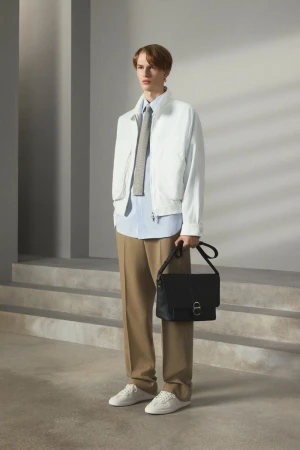 Male fashion model poses indoors wearing a white jacket, striped shirt, tan pants, white sneakers, and carrying a black bag.