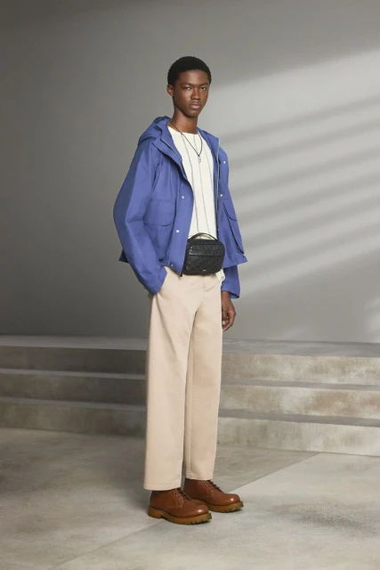 Fashionable adult man wearing a blue hooded jacket with beige trousers, and brown leather boots stands indoors in soft light.