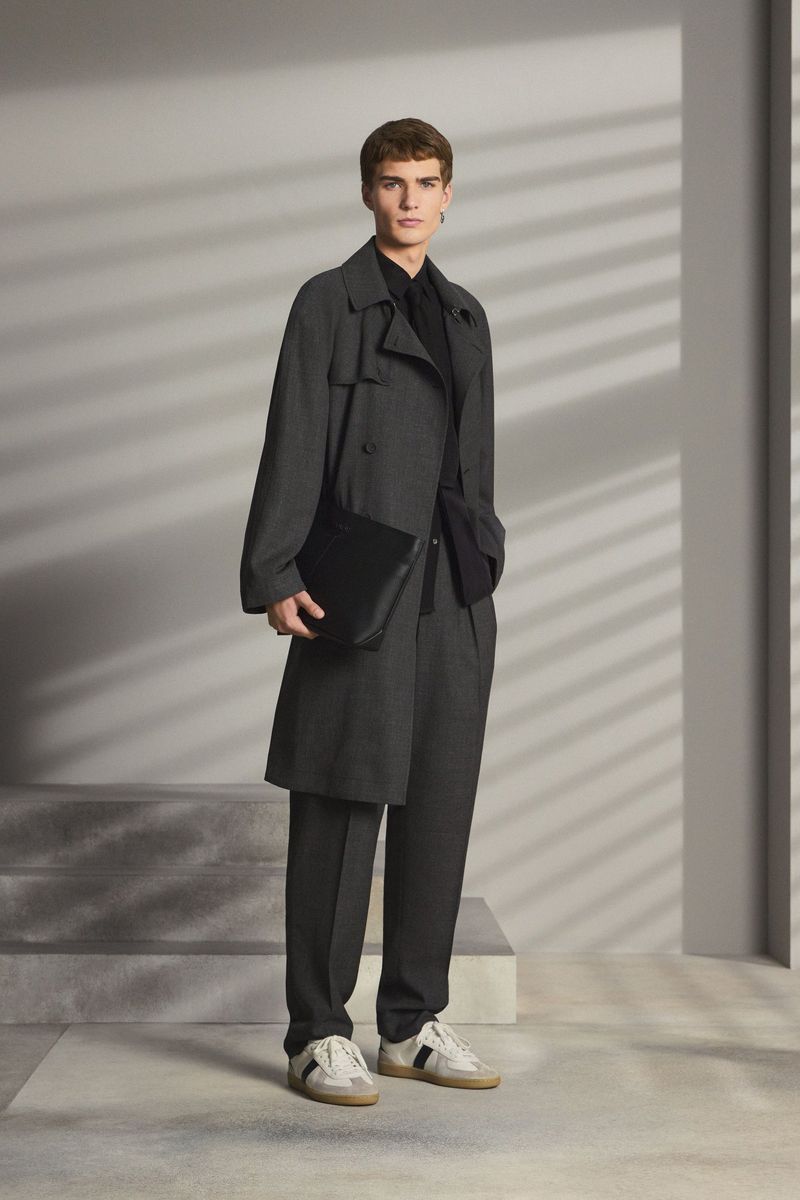 Male fashion model posing in a grey overcoat, black shirt and trousers, paired with sneakers and a black leather messenger bag.