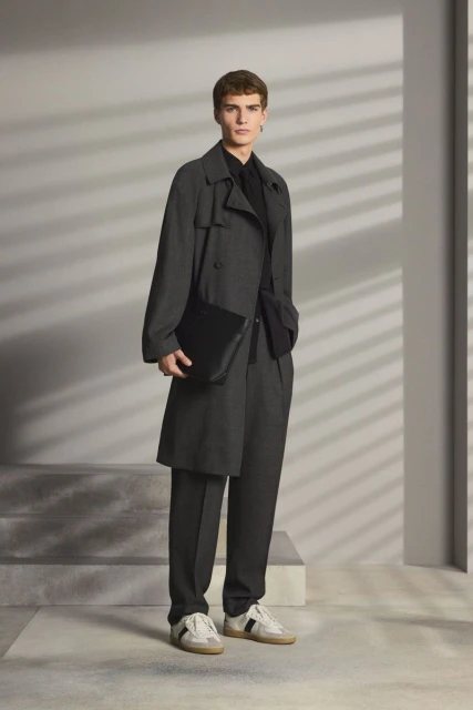 Male fashion model posing in a grey overcoat, black shirt and trousers, paired with sneakers and a black leather messenger bag.