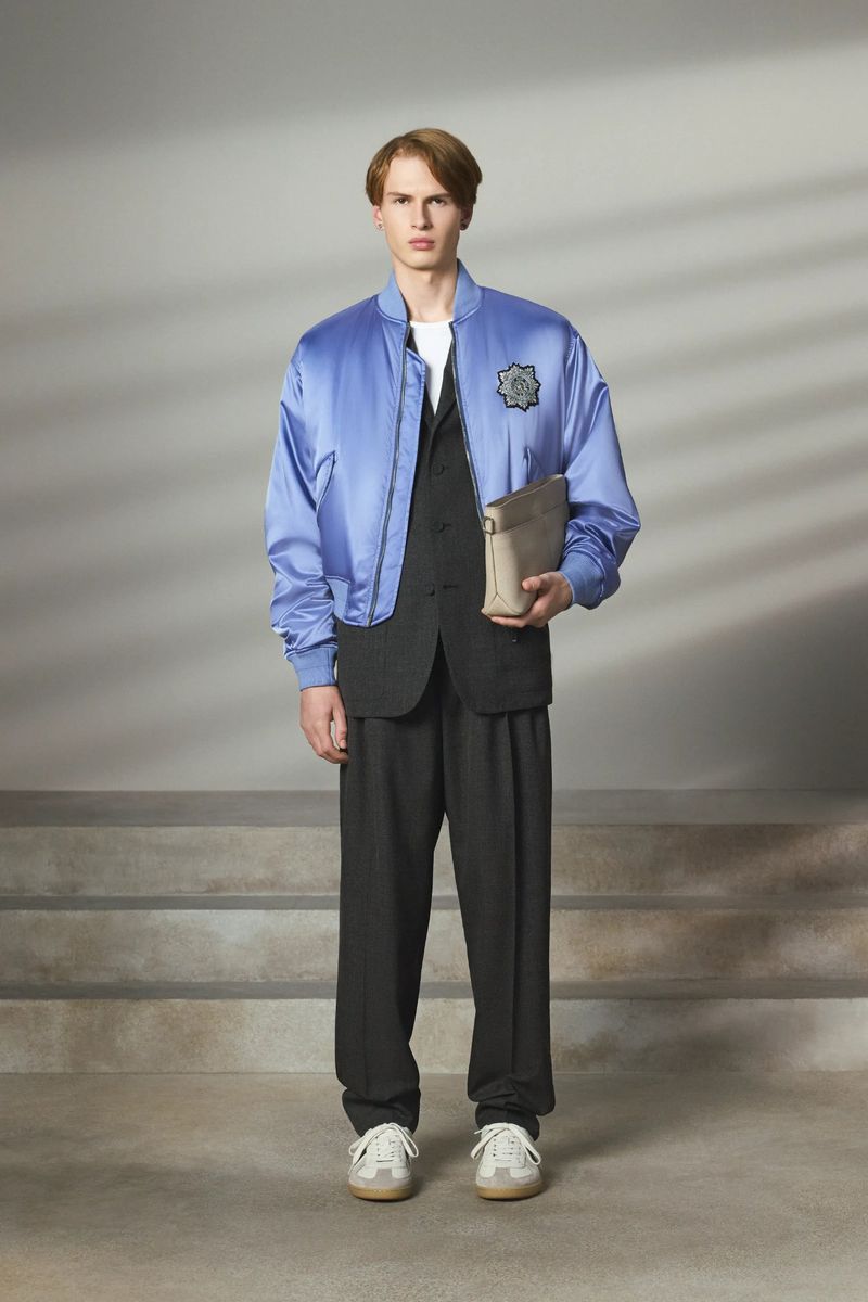 Male fashion model poses in a blue bomber jacket, black vest and trousers, with cream sneakers in a studio with soft lighting.
