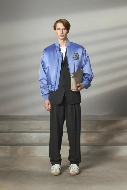 Male fashion model poses in a blue bomber jacket, black vest and trousers, with cream sneakers in a studio with soft lighting.