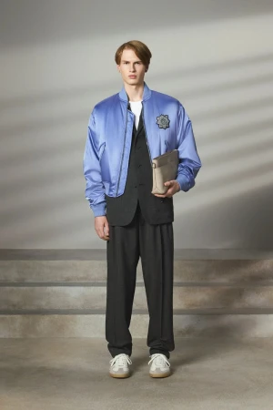 Male fashion model poses in a blue bomber jacket, black vest and trousers, with cream sneakers in a studio with soft lighting.