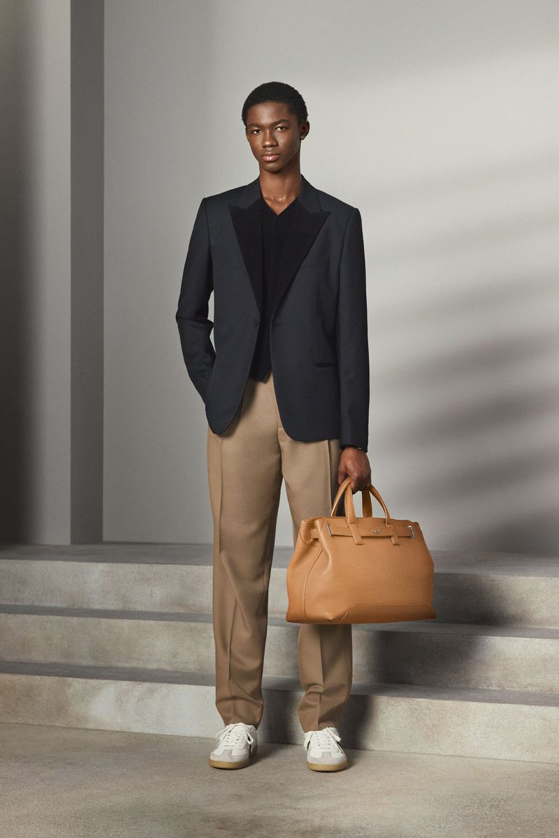 A young Black man posing on concrete steps wearing a dark blazer, V-neck top, camel trousers, and holding a large tan leather handbag.