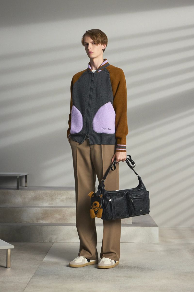 A young, slender man models knitwear and a black leather bag with teddy bear charm, indoors in front of a light grey wall.