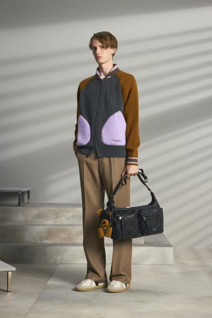 A young, slender man models knitwear and a black leather bag with teddy bear charm, indoors in front of a light grey wall.