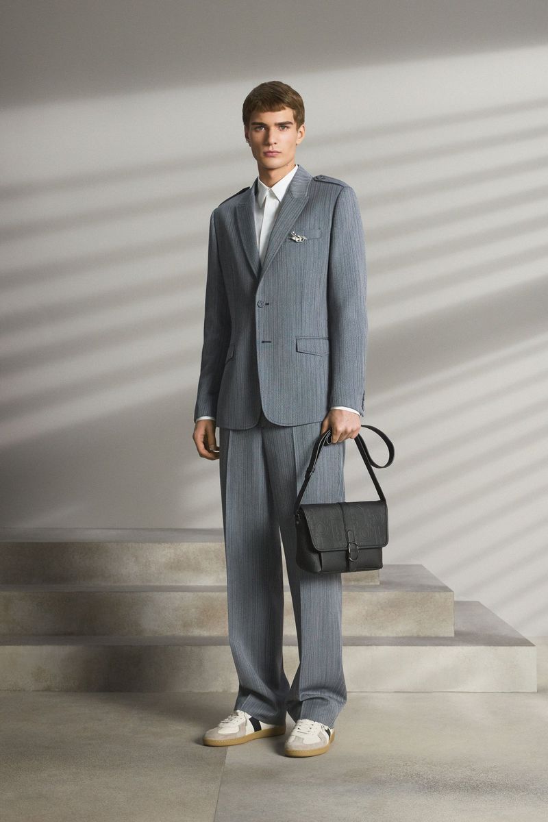 Full-length studio shot of a man in a gray pinstripe suit holding a black leather bag. He wears white trainers, in front of stairs.