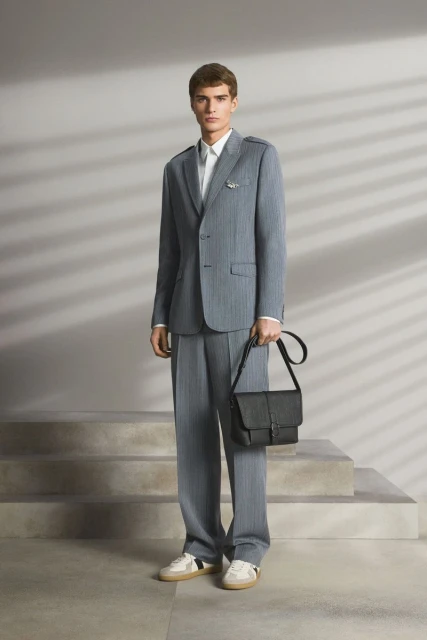 Full-length studio shot of a man in a gray pinstripe suit holding a black leather bag. He wears white trainers, in front of stairs.