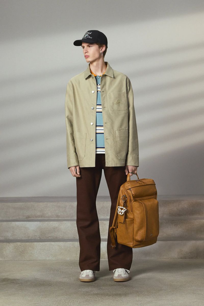 Man in beige jacket and brown pants poses with brown leather backpack against a gray background with subtle shadow pattern and steps.