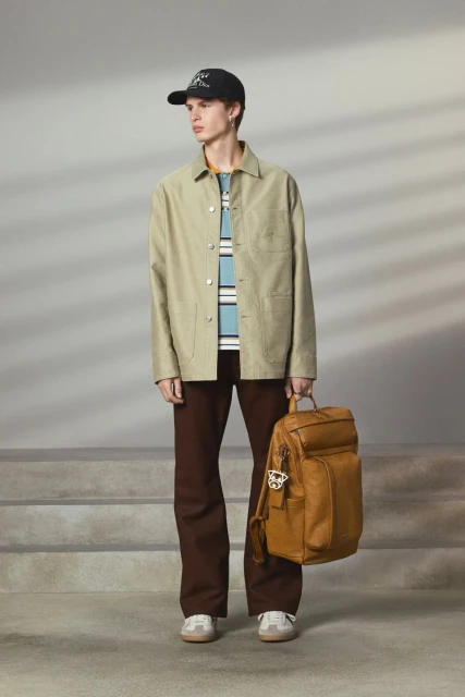 Man in beige jacket and brown pants poses with brown leather backpack against a gray background with subtle shadow pattern and steps.