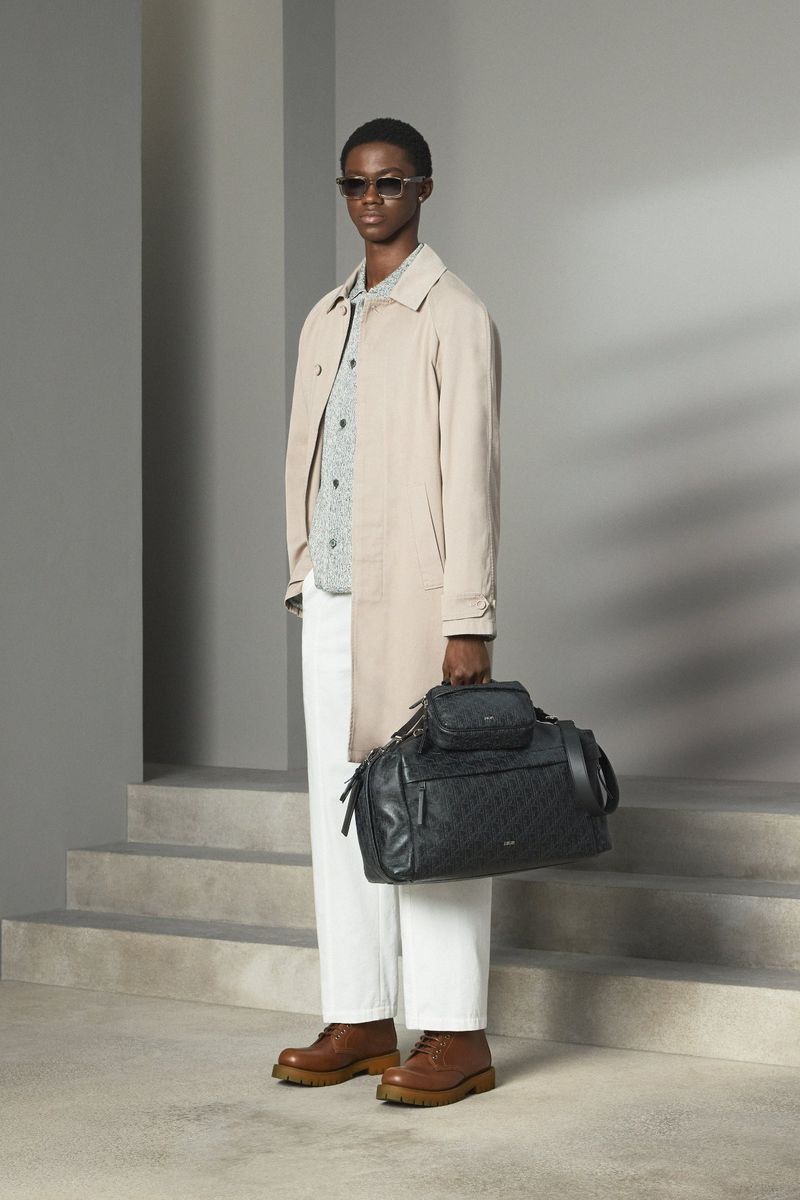 An adult male model stands in a studio wearing a trench coat, patterned shirt, white trousers, and brown boots holding a black travel bag.