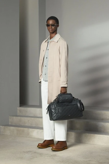 An adult male model stands in a studio wearing a trench coat, patterned shirt, white trousers, and brown boots holding a black travel bag.