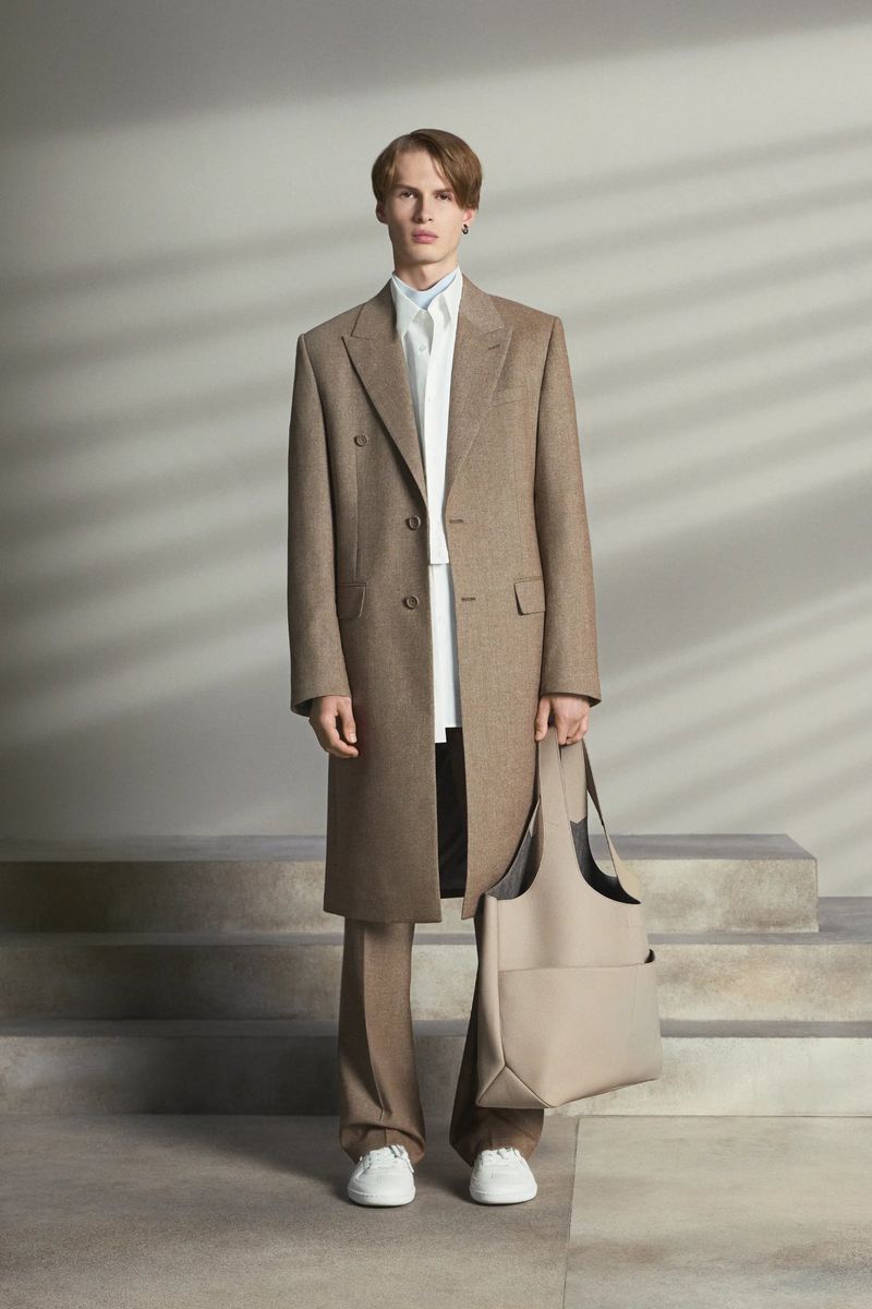 A young adult man stands in a studio wearing a brown overcoat, white shirt, and tan pants. He is holding a beige tote bag.