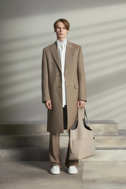 A young adult man stands in a studio wearing a brown overcoat, white shirt, and tan pants. He is holding a beige tote bag.