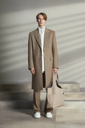 A young adult man stands in a studio wearing a brown overcoat, white shirt, and tan pants. He is holding a beige tote bag.