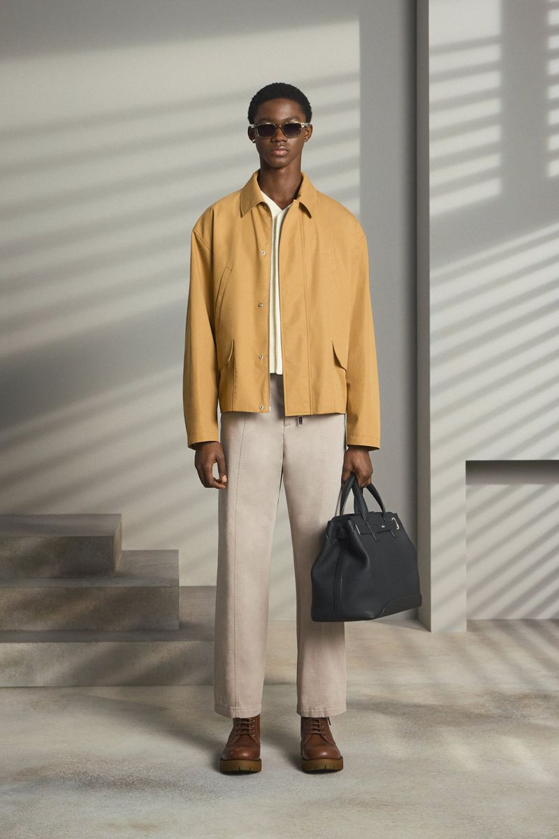 Adult male fashion model poses in a tan jacket and slacks holding a black leather tote bag in a neutral indoor studio setting.