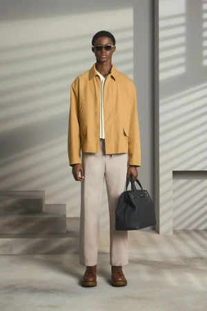 Adult male fashion model poses in a tan jacket and slacks holding a black leather tote bag in a neutral indoor studio setting.