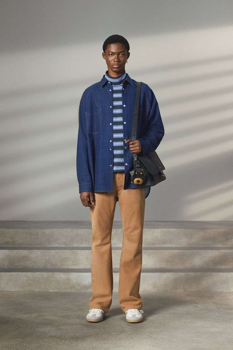 Full-length shot of a Black male fashion model wearing a blue denim shirt jacket, striped shirt, khaki pants, sneakers, and shoulder bag.